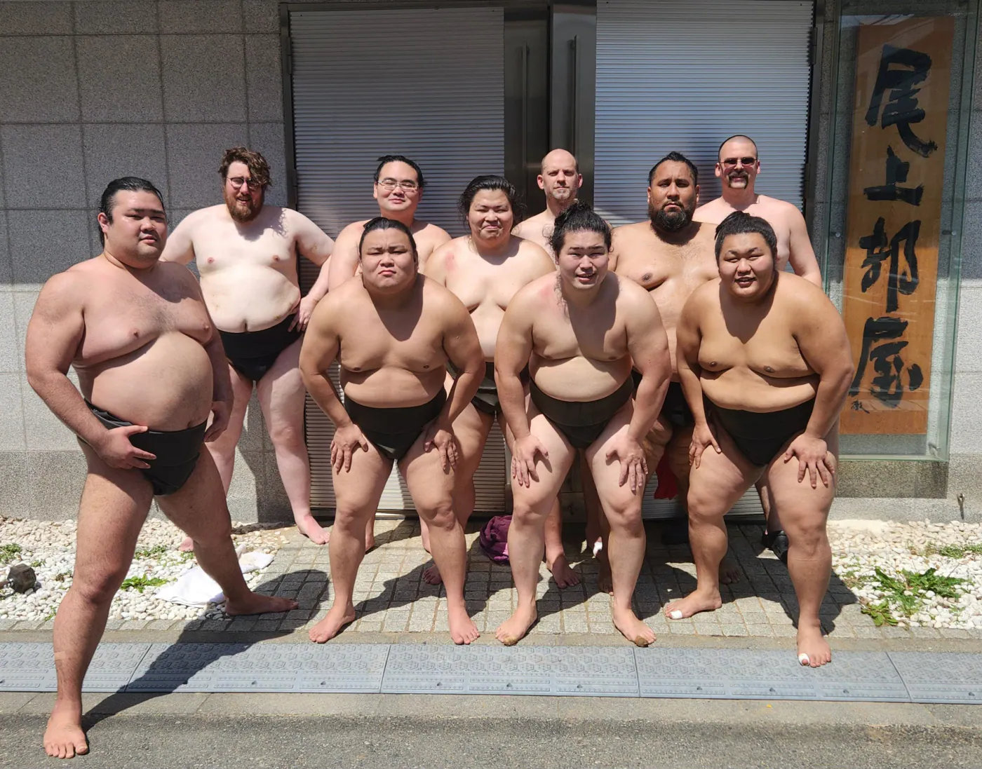 Wrestlers posing outside a sumo stable beside a calligraphy sign